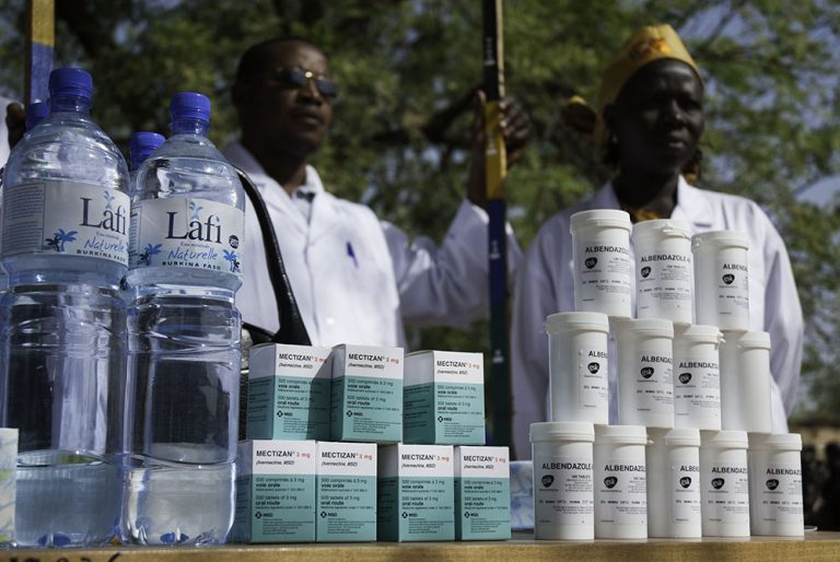Photo of men standing behind bottles of Mectizan, albendazole, and water