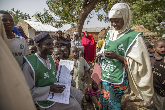 A man sits with a registration book while a woman dispenses tablets from a bottle. They are standing outdoors and are surrounded by children and adults who are waiting to receive medicine.