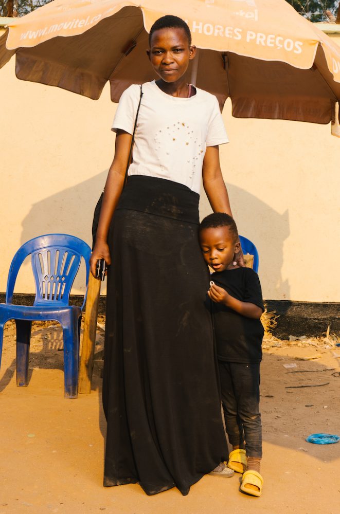 A woman wearing a long black skirts stands with her little boy in an open-air market.