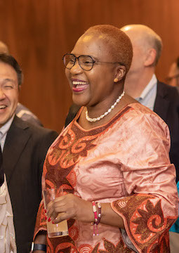 Photo of a woman who is laughing while standing in a group of people at a formal event. She is wearing pearls and a traditional dress with an orange pattern.
